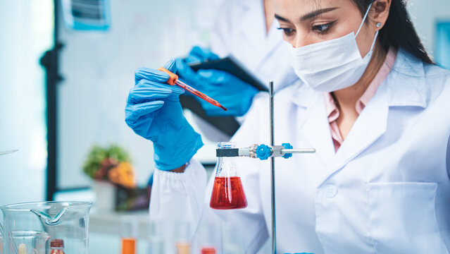 A gloved scientist carefully drips red solution into a flask, with red test tubes in the foreground, symbolizing precision, collaboration, and progress in modern medical dna blood research. - Powered by Adobe