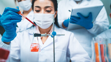 A female scientist drops a blood sample into an Erlenmeyer flask while colleagues record data,...