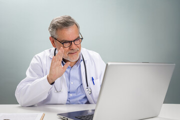 A doctor in a white coat is smiling and waving at the camera during an online consultation. The setting is well-lit and equipped with a laptop, creating a friendly virtual environment.
