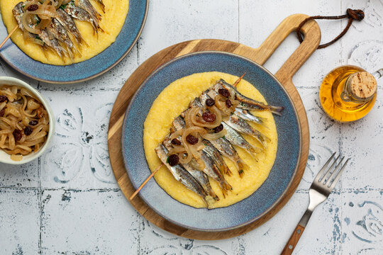 Italian meal. Polenta with grilled Sardines skewers and saor, a traditional Venetian sweet and sour marinade of onions, raisins. Mediterranean food in a blue plate on a light table surface.