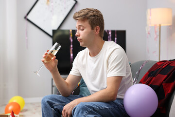 Man with glass of alcohol suffering from hangover on sofa in messy room after party