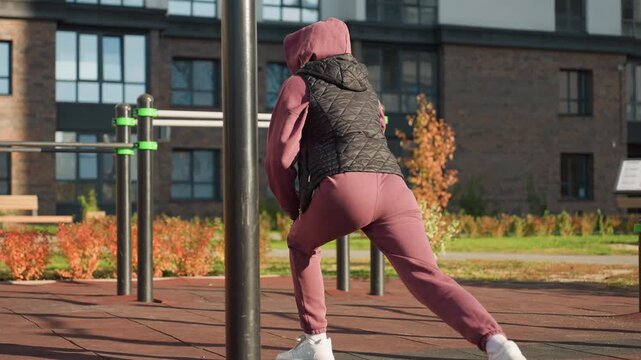 Back view of active woman in sports wear doing split squats with hands placed on legs in modern urban park environment featuring flowers rails shadows on rubber tile under bright sunlight