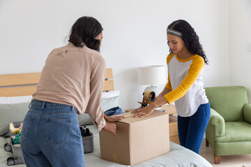 Diverse mom and daughter sealing cardboard box with tape dispenser near folded clothes on bed