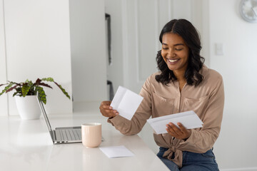Smiling woman with envelopes and letters at kitchen countertop with laptop mug plant
