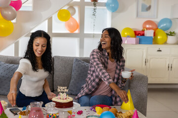 Diverse mom and teenage daughter sitting on couch in living room preparing cake balloons hats
