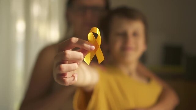 Emotional moment of mother hugging her teenage son who holds yellow satin ribbon symbolizing pediatric cancer awareness and hope.