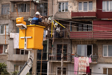An electrician in a hard hat in a yellow aerial work platform repairs city lighting after broken wires on utility poles in Georgia, with a multi-story building in the background.
