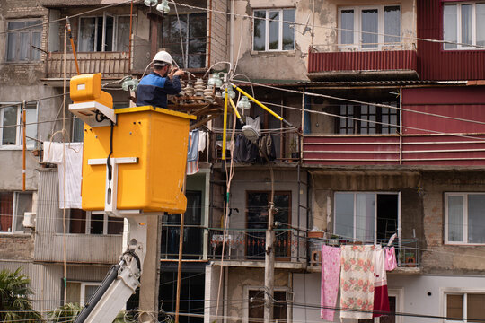 An electrician in a hard hat in a yellow aerial work platform repairs city lighting after broken wires on utility poles in Georgia, with a multi-story building in the background.