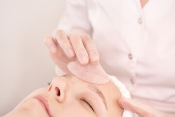 Fototapeta premium Young caucasian female receiving facial treatment with rose quartz gua sha in spa.