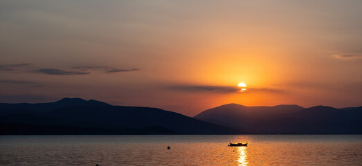 Tranquil fishing boat at sunset over Tolo Bay Greece. Glowing orange sky and distant mountain silhouettes reflecting on calm sea water
