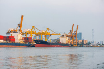 Large cargo ships docked under towering yellow container cranes loading shipping containers with flat barge and tugboat navigate on Chao Phraya River in Bangkok, Thailand. Maritime and logistics hub