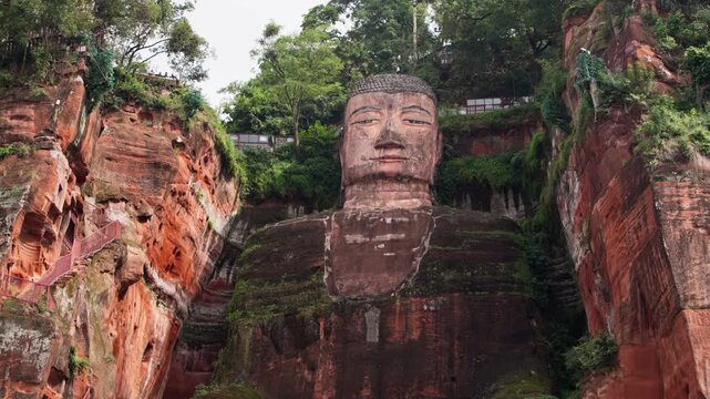 Cinematic slow motion pan across the top of the Leshan Giant Buddha, with rich red rock and surrounding greenery