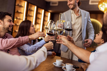 Group of young friends in modern cafe raising glasses in toast while one man stands, cheerful social moment.