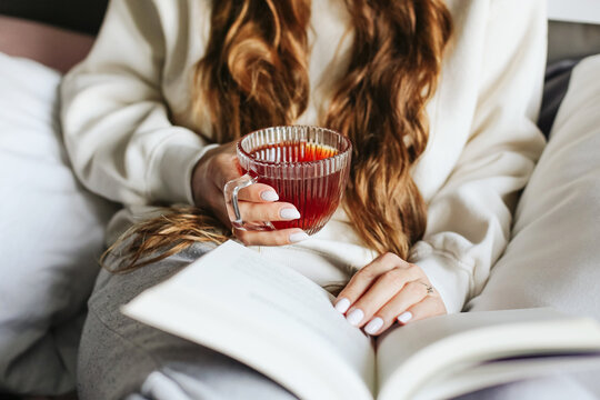 Woman holding glass cup of tea and reading book. Cozy lifestyle moment with warm drink. Female hands closeup with long hair. Relaxing leisure time at home. Peaceful evening atmosphere indoors.