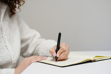 A woman in a white shirt is writing in a notebook with a black pen on a clean white desk
