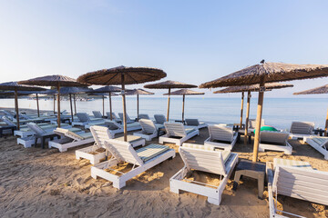 Well‑kept beachfront lined with white wooden lounge chairs and straw umbrellas on soft sand, facing calm blue waters under a clear sky, promoting coastal getaways, relaxation, and summer holidays.