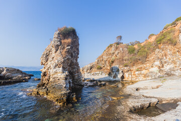 Towering limestone sea stack rising from crystal clear Mediterranean waters with layered sedimentary cliffs and rocky coastline under bright blue sky.