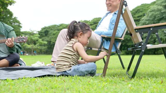 Happy multi generational Asian family enjoying picnic in the park, Grandfather playing guitar creating joyful outdoor lifestyle moment