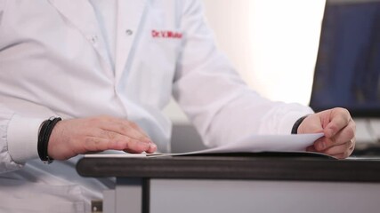 A doctor wearing a white lab coat sits at a desk, reviewing documents and speaking to someone. The scene suggests a consultation, diagnosis - Powered by Adobe