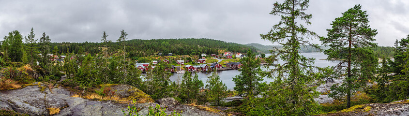 Panoramic view of Bonhamn, a charming fishing village on the Swedish coast, featuring red huts along the water's edge, surrounded by lush pine trees and dramatic clouds