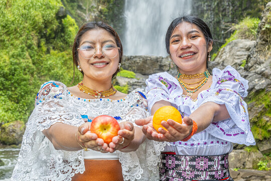Two Otavalo indigenous women in traditional attire hold fresh fruits, a red apple and an orange, in front of the Peguche waterfall, Ecuador. Reflecting connection with nature and healthy living.