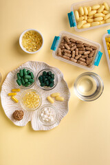 Vitamins, minerals and dietary supplements in containers and glass bowls, glass of water from above on yellow background. Assortments of many colored pills, tablets, capsules and food supplements. 