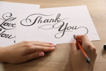 Calligraphy. Woman with brush writing words Thank You on sheet of paper at wooden table, closeup
