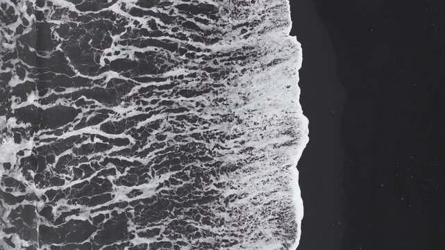 Top down aerial view of a black sand beach in Iceland, showing waves crashing and retreating rhythmically against the volcanic shoreline.