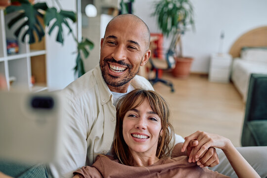 Happy multi-ethnic couple enjoying a cozy moment together, taking a selfie on their smartphone while relaxing on the sofa at home - Powered by Adobe