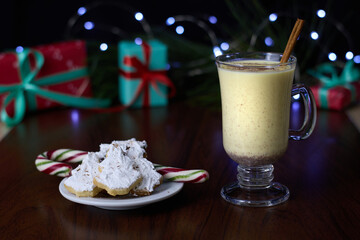 Eggnog served in a glass, accompanied by a cinnamon stick and Christmas tree-shaped cookies coated with white icing, set against a backdrop of a pine branch with twinkling fairy lights.