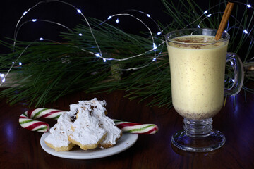 Eggnog served in a glass, accompanied by a cinnamon stick and Christmas tree-shaped cookies coated...