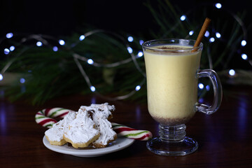 Eggnog served in a glass, accompanied by a cinnamon stick and Christmas tree-shaped cookies coated with white icing, set against a backdrop of a pine branch with twinkling fairy lights.