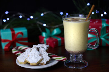 Eggnog served in a glass, accompanied by a cinnamon stick and Christmas tree-shaped cookies coated with white icing, set against a backdrop of a pine branch with twinkling fairy lights.