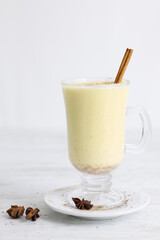 A festive arrangement featuring eggnog, cinnamon sticks, star anise, a candy cane, and Christmas tree-shaped cookies coated with white icing, all placed on a gray table.