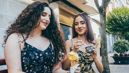 Two young friends, both in summer dresses and sunglasses, pose with ice cream in the street. One...
