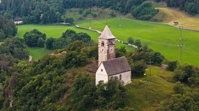 Chiesa di Santa Verena on a gentle hilltop with fields below, aerial view. Dolomites landscape with agricultural parcels