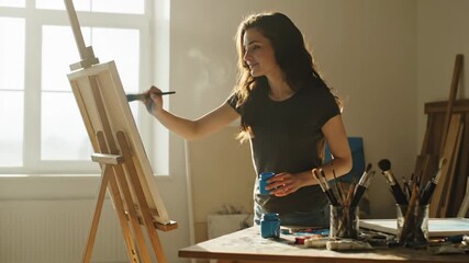 Young Artist Painting on Canvas in Studio - A young woman with long brown hair paints on a canvas easel in her bright art studio. - Powered by Adobe