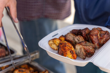 A hand uses tongs to place grilled meat into a foam takeout container at a barbecue