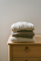 Stack of folded warm knitted sweaters in beige, brown and white tones placed on a dresser. Cozy autumn and winter mood.