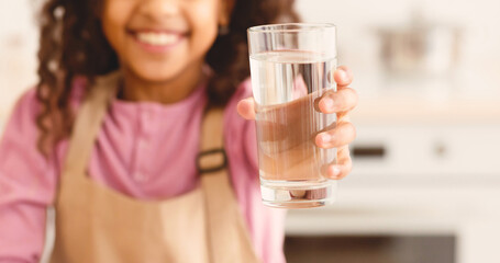 Closeup Shot Of Cute Little Black Girl Holding Glass Of Water In Hand And Giving It At Camera, Enjoying Drinking Healthy Refreshing Beverage, Sitting At Table In Kitchen At Home, Selective Focus