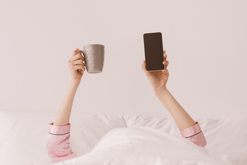 Hands of millennial caucasian lady in pink pajamas stick out from white blanket and hold cup of...