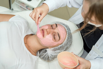 Obraz premium Beautiful woman receiving ping clay facial mask by beautician at spa salon