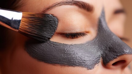 Woman with eyes closed, receiving a facial treatment as a brush applies a gray clay mask