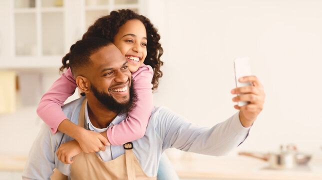 Fatherhood Concept. Portrait of cheerful african american father taking selfie with his little daughter while cooking in the kitchen together at home, giving her piggyback ride, using smartphone - Powered by Adobe