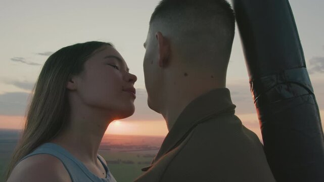 close up young couple kissing and gazing at vibrant sunset horizon from hot air balloon basket as gentle breeze rustles hair and distant paraglider drifts over sprawling green fields under pastel sky