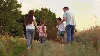 Loving family with little kids and bicycle walks along rural road at sunset backside view. Father helps boy to ride bicycle mother and daughter walk joining hands on family country vacation in summer