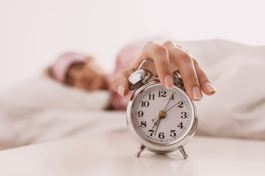 Lady lying on bed and trying wake up, get up early for work. Sleepy young blonde woman in pink pajamas wake up and turns off alarm clock on white soft comfortable bed in bedroom in morning, blurred
