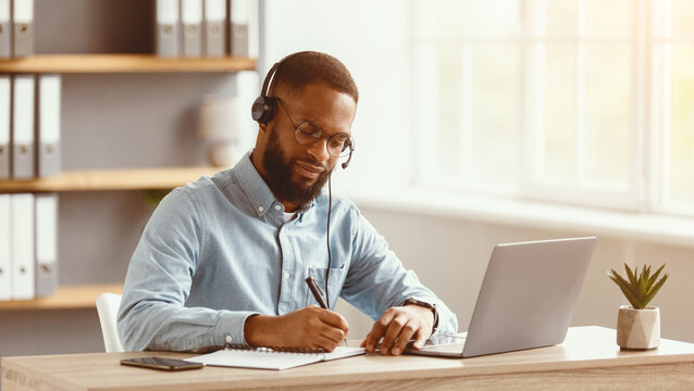 Busy serious millennial african american bearded guy in glasses and headset working on laptop in home office interior. Customer support, remote job, business indoor and social distancing, free space