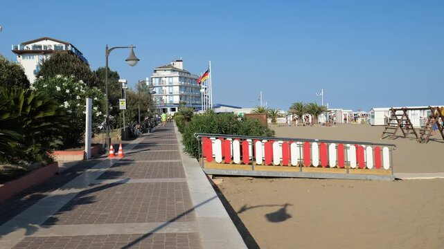 A tourist walks along the promenade near a large seaside beach where many people are relaxing. Summer heat in Italy