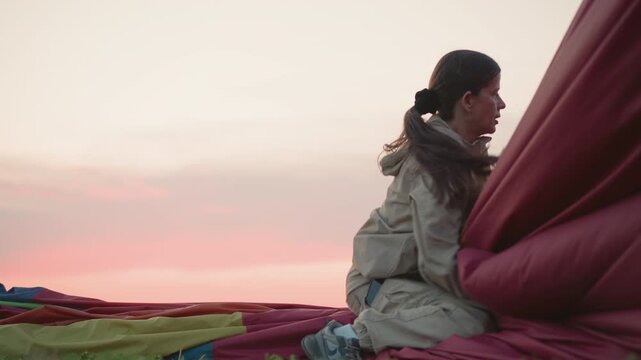 woman kneeling on deflated hot air balloon fabric on green field at sunset grasping red envelope panel under soft pastel sky during twilight countryside landing pack up operation
